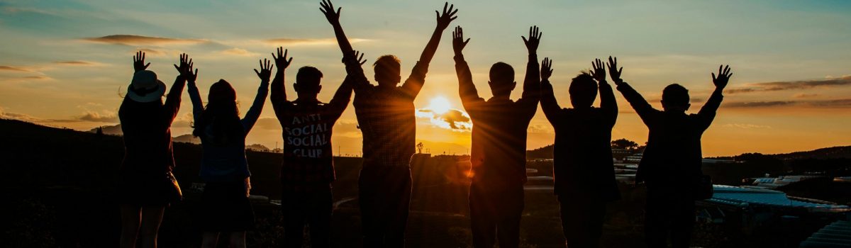 A diverse group of friends raises their arms in celebration against a vibrant sunset backdrop.